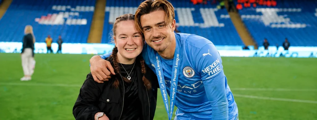 Hollie Grealish smiling and posing alongside her older brother, Manchester City footballer Jack Grealish, on the pitch during a Premier League title victory celebration at the Etihad Stadium.